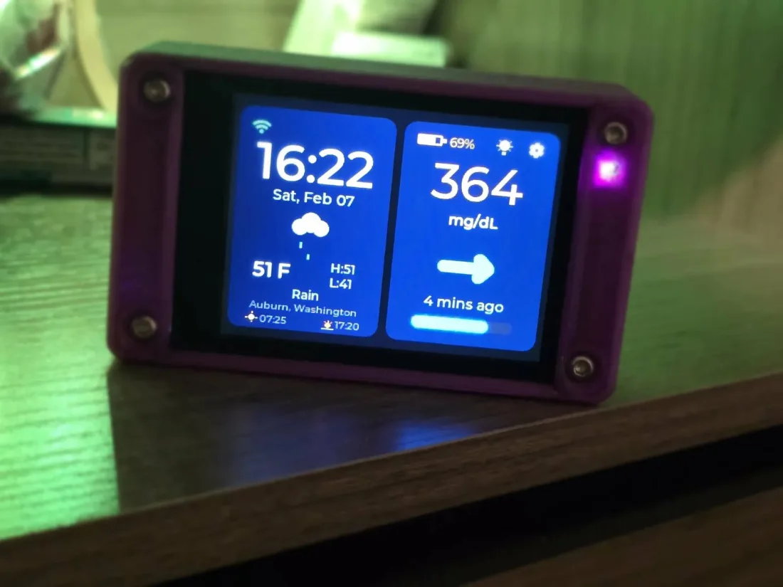 A CYGM unit in a purple 3D-printed case on a desk, its screen showing the time, local weather and a glucose reading with a trend arrow